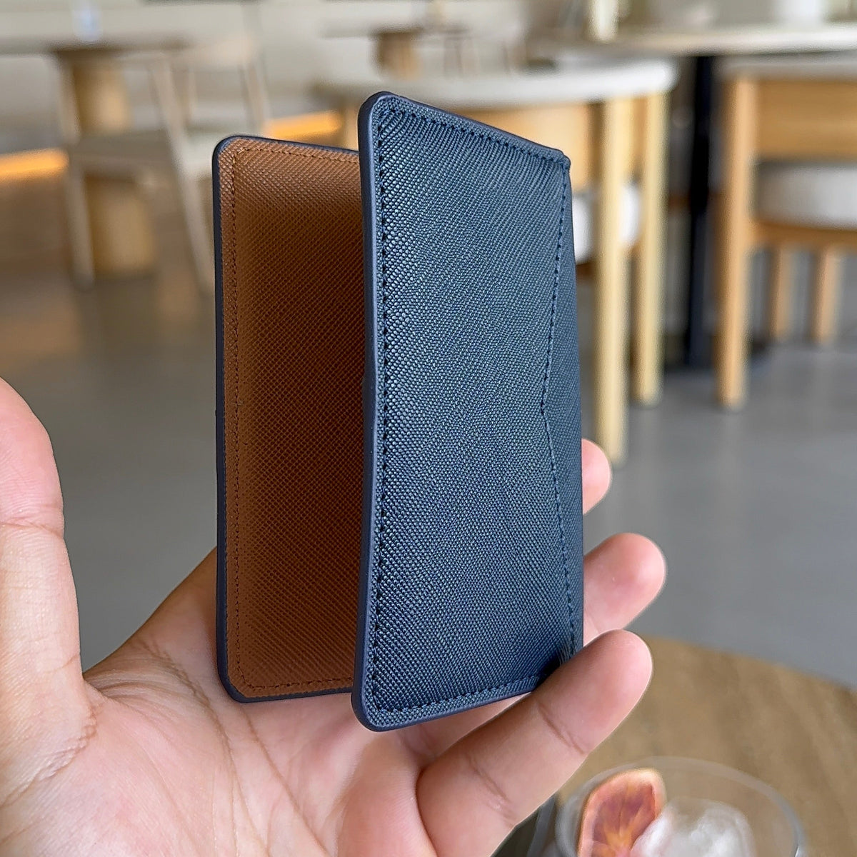 A hand holding a slim two-tone card holder or wallet with a dark blue textured exterior and a brown interior, shown in a blurred cafe setting.